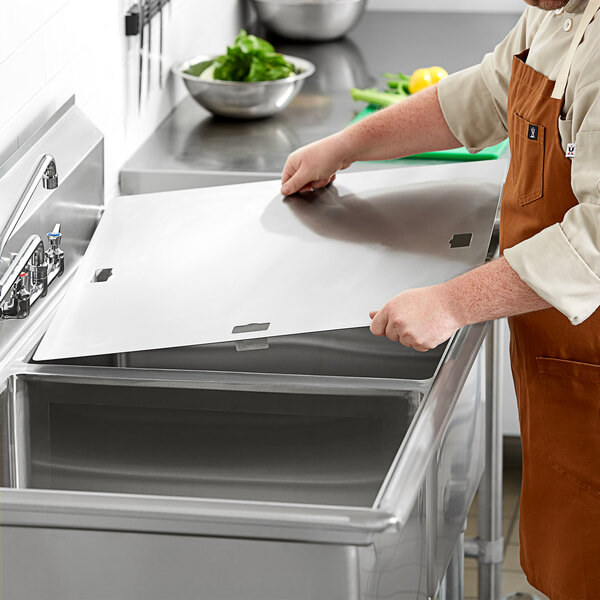 A man in an apron opening a stainless steel sink cover over a commercial sink.