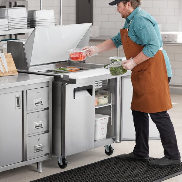 An aproned man using an Avantco mega top sandwich prep table to put food in a container.