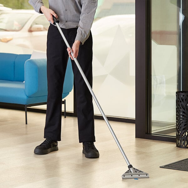 A man using a Lavex heavy-duty floor scraper with a long handle to clean a floor in a professional kitchen.