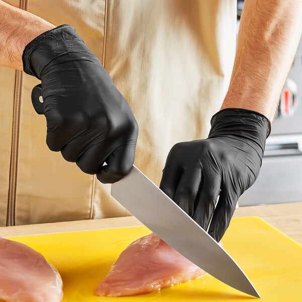 A person wearing black Noble NexGen biodegradable nitrile gloves cutting a piece of meat on a cutting board.