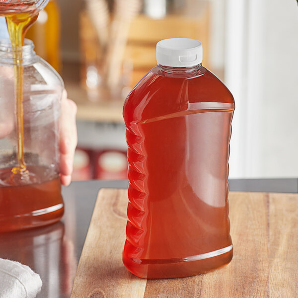 A person pouring honey into a Ribbed Hourglass PET Honey Bottle on a cutting board.