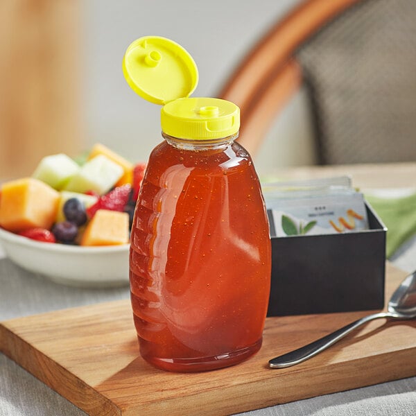 A Classic Queenline PET honey bottle with a yellow plastic flip top lid on a cutting board with a bowl of fruit.