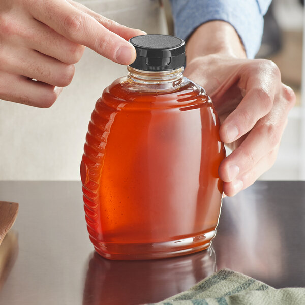 A person holding a 16 oz. Queenline PET honey bottle with a black plastic flip top lid full of honey.