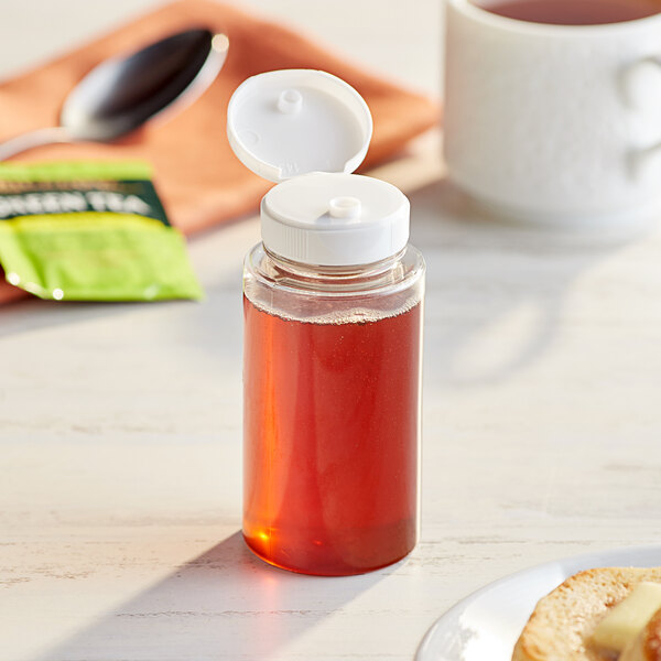A 5.5 oz. clear PET sauce bottle with a white cap on a table with a jar of honey and a cup.