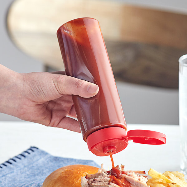 A hand pouring from a clear sauce bottle with a red lid onto a sandwich.
