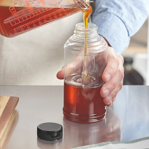 A hand pouring brown liquid into a Skep sauce/honey bottle with a black flip top lid.