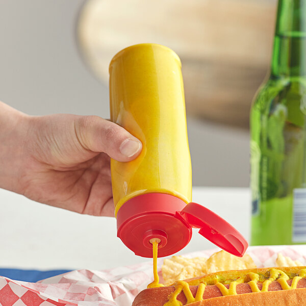 A hand holding a yellow bottle of mustard squeezes mustard onto a hot dog.