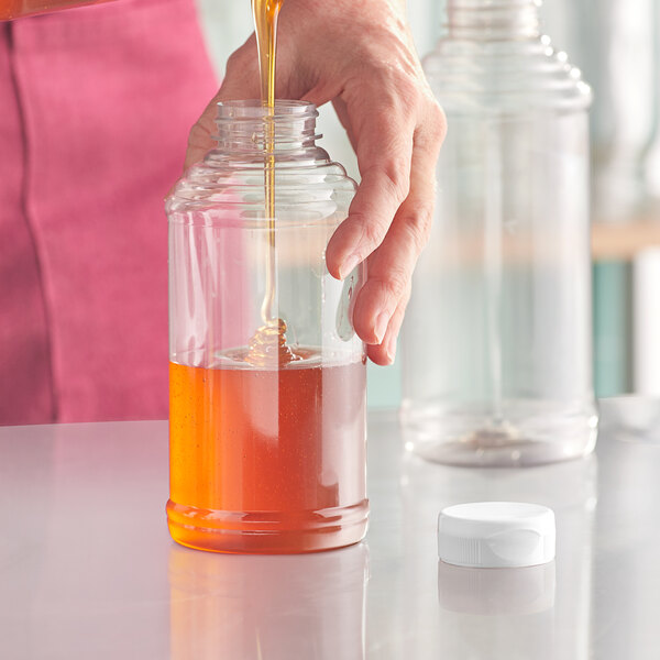 A person pouring honey from a white bottle into a glass jar.