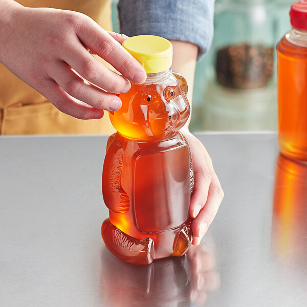 A person pouring honey into a bear-shaped PET honey bottle with a yellow cap.