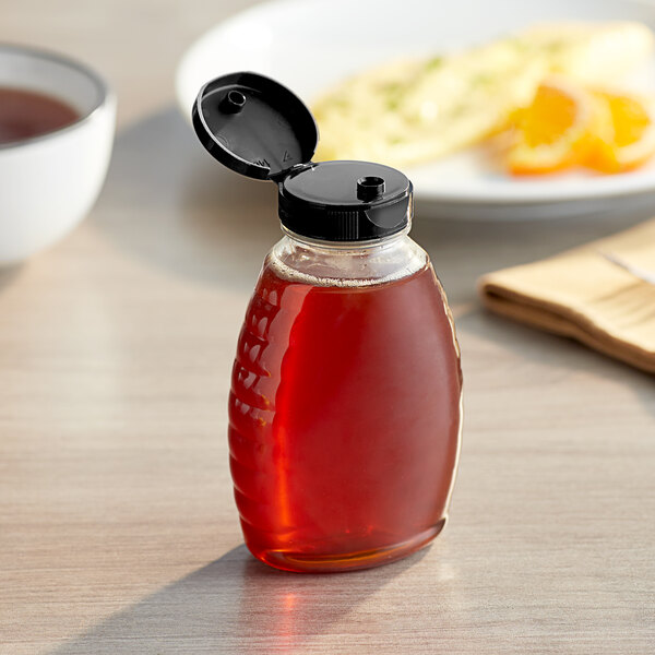 A Classic Queenline PET honey bottle with a black cap on a table next to food.