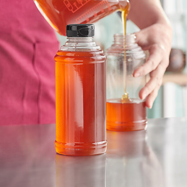 A woman pouring honey from a bottle with a black cap into a glass jar.