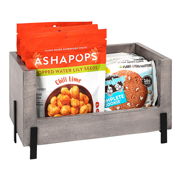 A Cal-Mil gray pine stacking box on a counter filled with bags of snacks.