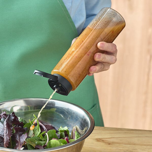 A person pouring sauce from a clear cylinder bottle with a black squeeze lid onto a salad.