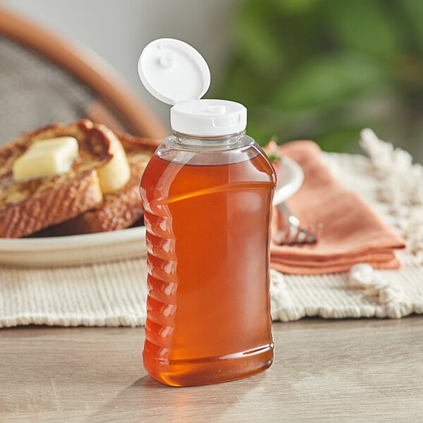 An 11 oz. ribbed hourglass PET honey bottle with a white plastic flip top lid filled with honey on a table next to toast.