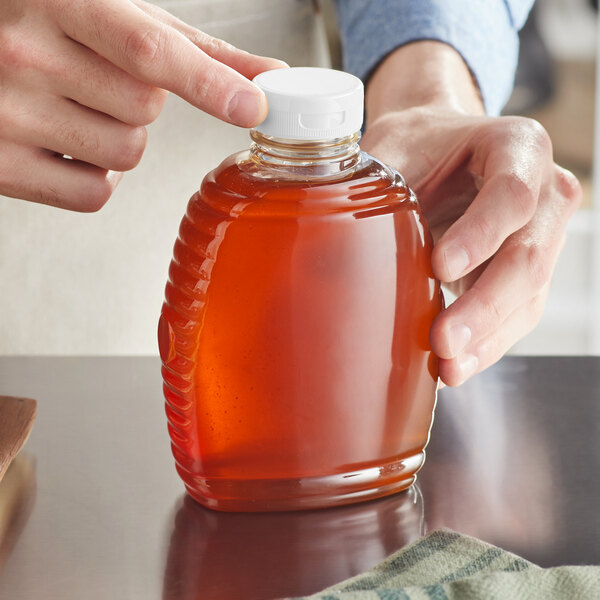 A person pouring honey from a Queenline PET Honey Bottle with a white flip top lid into a jar.