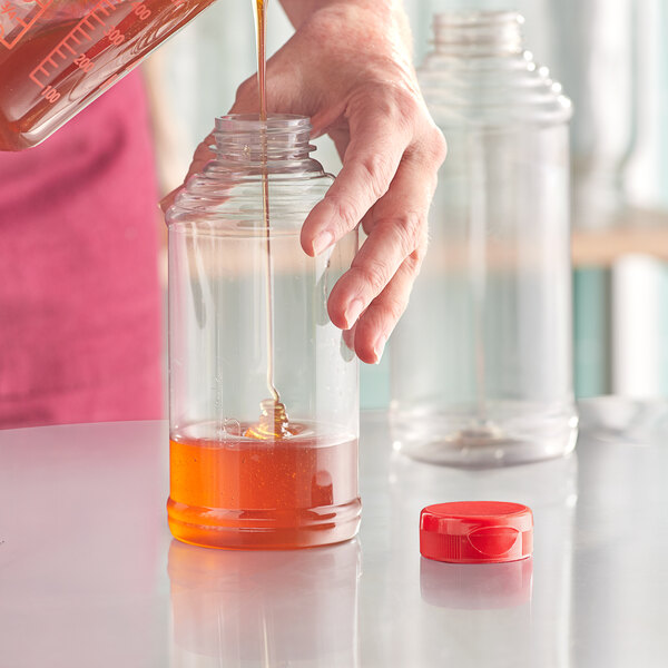 A person pouring honey into a 16 oz. Skep PET sauce bottle with a red cap.