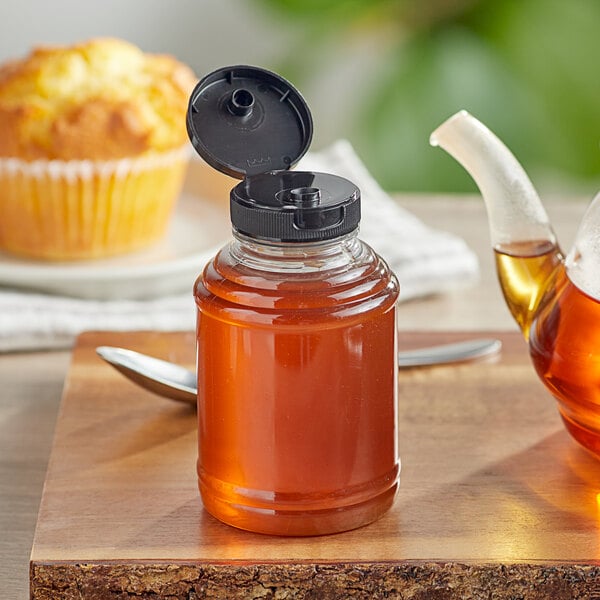 A jar of honey with a black flip top lid next to a muffin on a table.