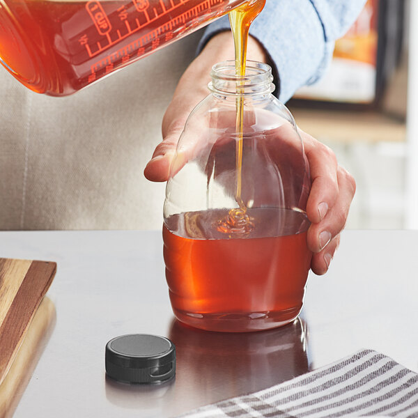 A person pouring honey into a Classic Queenline PET honey bottle with a black plastic flip top lid.