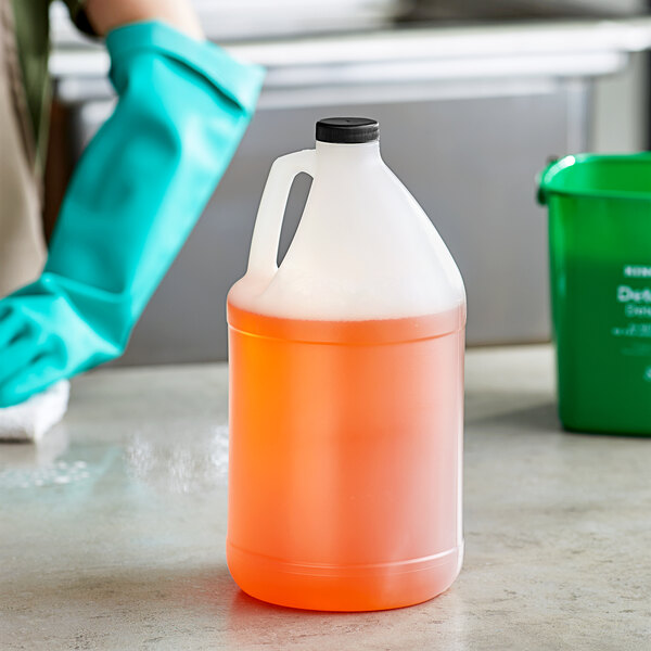 A gloved hand holding a translucent HDPE jug of orange liquid.