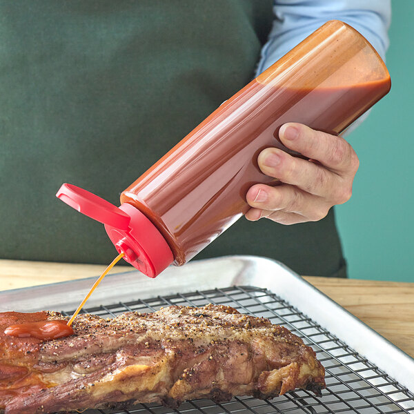 A person using a 16 oz. clear sauce bottle with a red squeeze lid to pour sauce on a piece of meat.