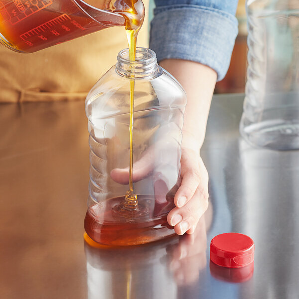 A person pouring honey into a Ribbed Hourglass PET Honey Bottle with a red cap.
