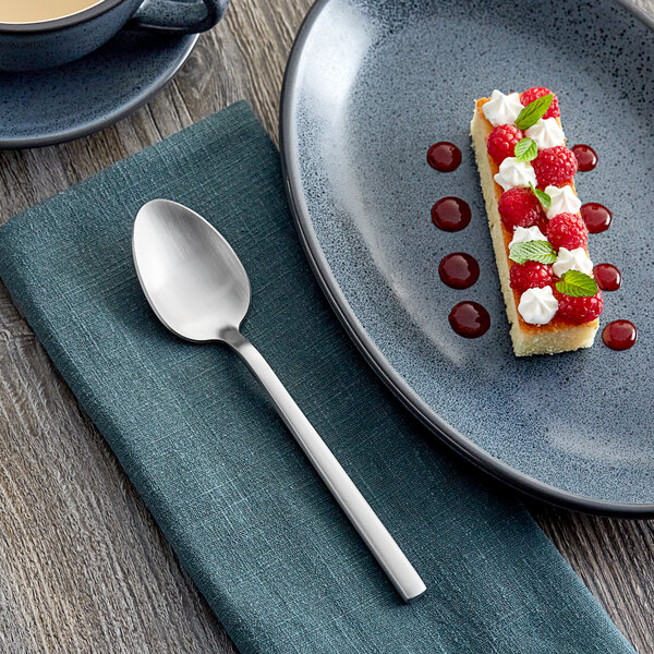 A plate with a piece of cake and an Acopa Phoenix stainless steel teaspoon on a napkin.