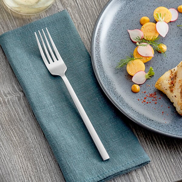 An Acopa stainless steel dinner fork on a napkin next to a plate of food.