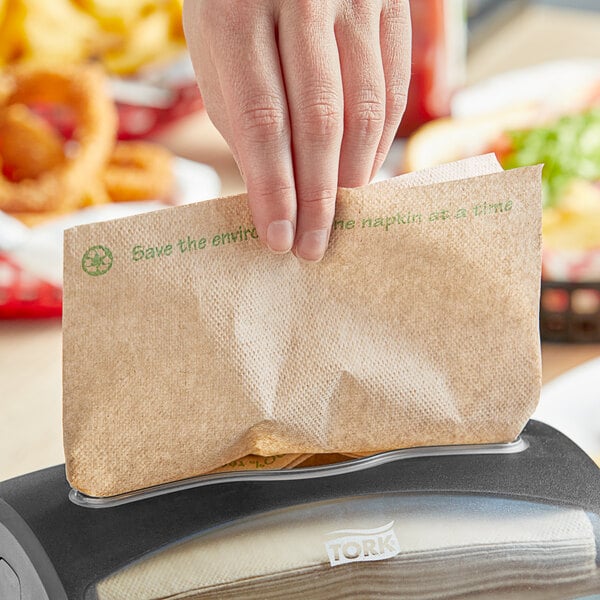 A hand holding a Tork Universal Xpressnap natural kraft dispenser napkin with green environmental print in front of a dispenser.