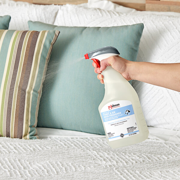 A hand holding a spray bottle of SC Johnson Professional Fabric and Air Deodorizer spraying a bed.