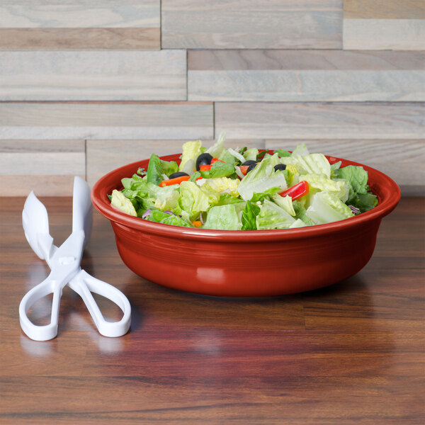 A Fiesta Scarlet china bowl filled with salad on a wood table with a pair of scissors.