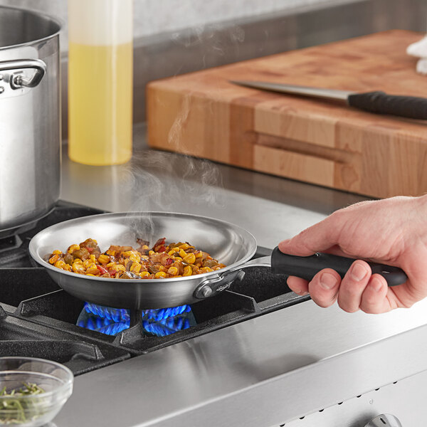 A hand using a Vollrath stainless steel fry pan with a black handle to cook food on a stove.