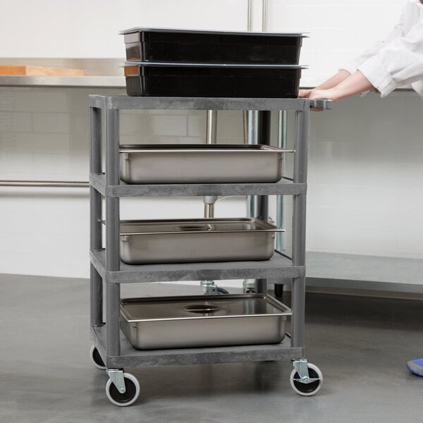 A person pushing a Luxor gray serving cart with metal trays.