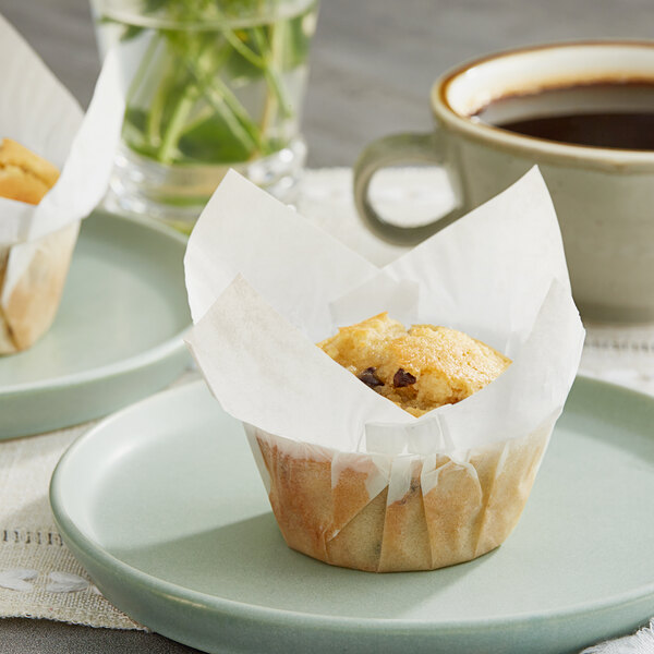 A plate with a muffin in a Baker's Mark white paper wrapper next to a cup of coffee.