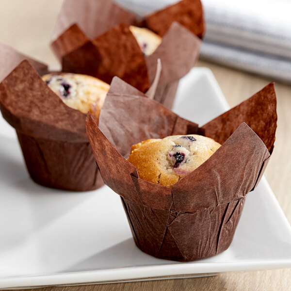 Three Baker's Mark chocolate brown muffins in large paper cups on a white plate.