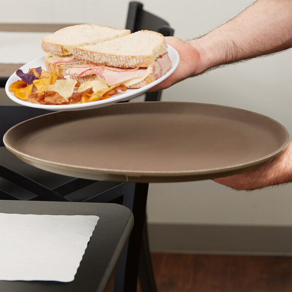 A hand holding a Carlisle brown non skid fiberglass serving tray with a sandwich with ham and cheese on it.