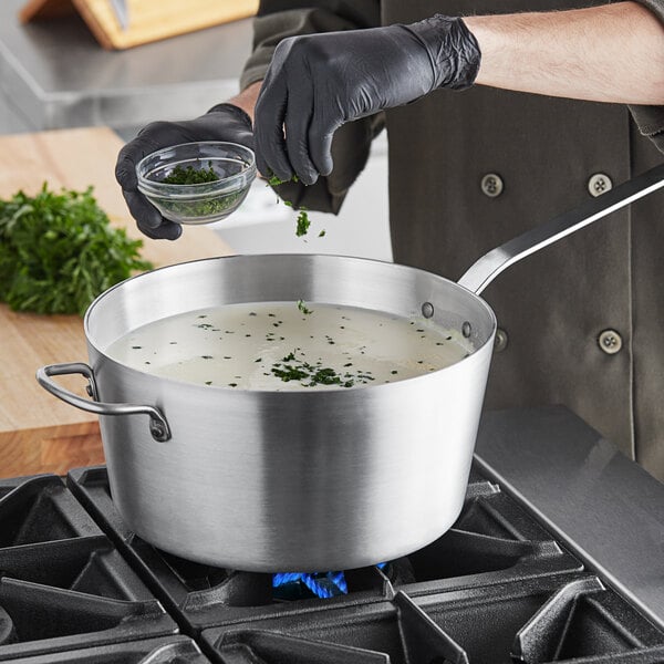 A person in black gloves pouring herbs into a Vollrath Wear-Ever sauce pan of soup on a stove.