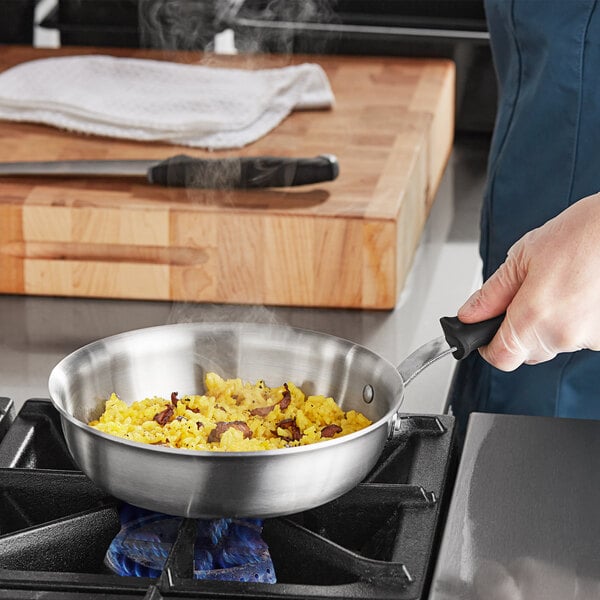 A person cooking food in a Vollrath stainless steel saucier pan on a stove.