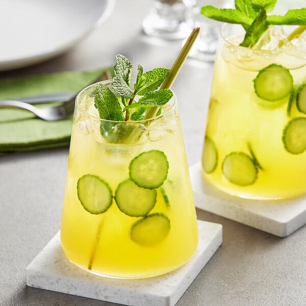 Two glasses of yellow liquid with cucumber slices on top on a table with a spoon and mint.