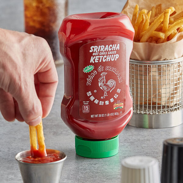 A person dipping a french fries into a red plastic container of Huy Fong Sriracha Hot Chili Ketchup.