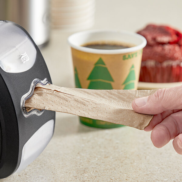 A hand holding a Tork Xpressnap Fit Advanced 2-Ply Natural Kraft interfold dispenser napkin next to a cup of coffee.