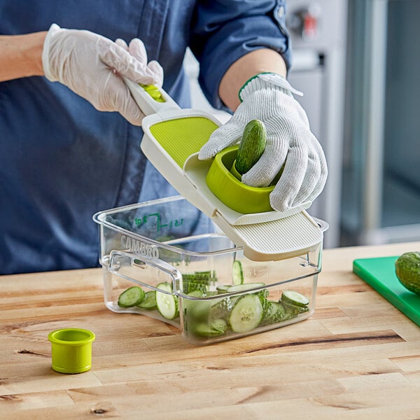 A person using a Choice Handheld Adjustable Mandoline to slice cucumbers into a green container.