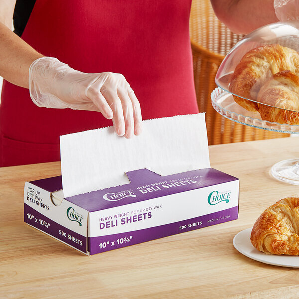 A person wearing a glove opening a purple and white box of Choice Interfolded Deli Wrap Paper to put a napkin in it.