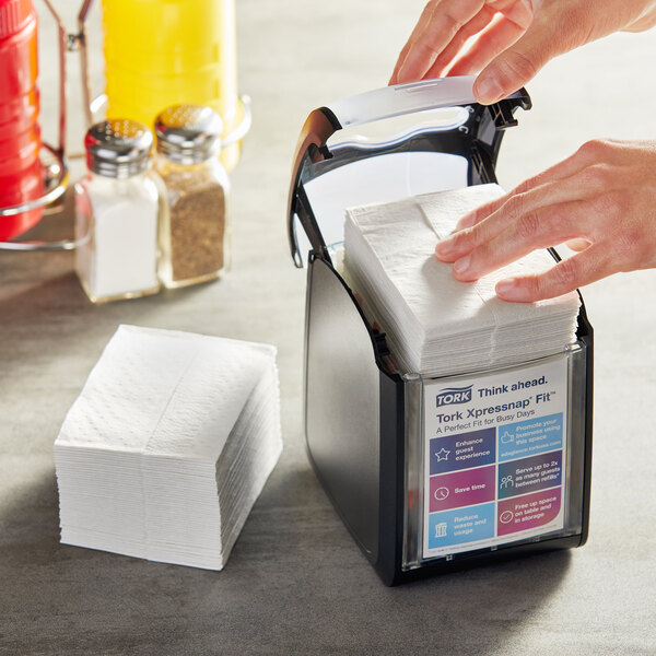 A person's hands using a Tork Xpressnap black tabletop napkin dispenser to take a napkin.