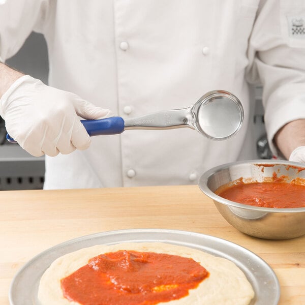 A person holding a Vollrath Blue Solid Round Spoodle over a pizza with red sauce.
