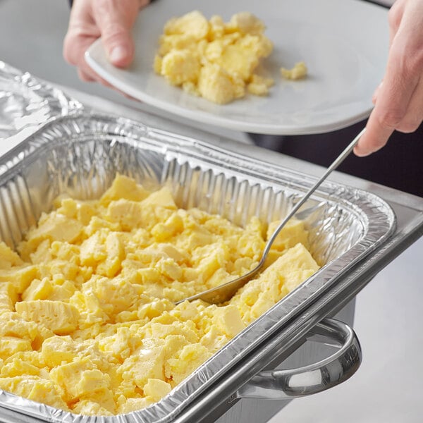 A person holding a metal spatula in a foil container of Grand Prairie scrambled eggs.