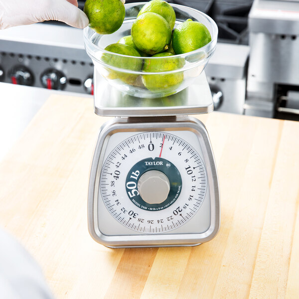 A hand holding a bowl of limes on a Taylor portion scale on a counter.