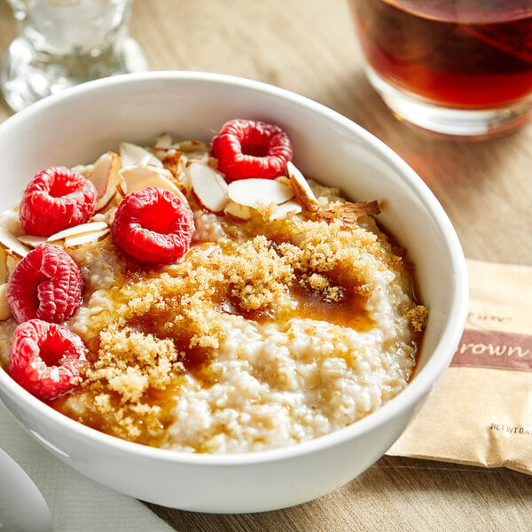 A bowl of oatmeal with raspberries and almonds and a brown sugar packet on a table.