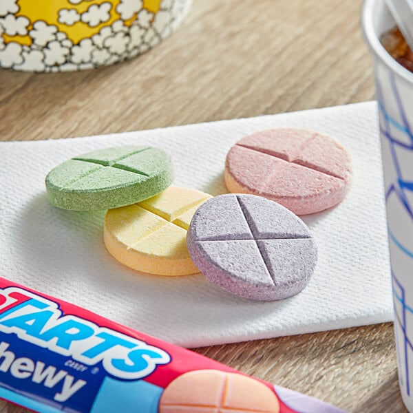 A box of SweeTarts Giant Chewy Candies on a table with a bowl of candy and a cup of soda.