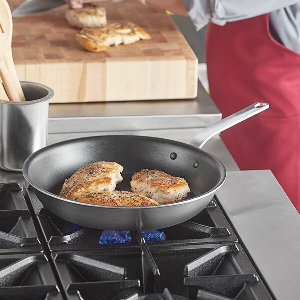 A person cooking chicken in a Vollrath stainless steel non-stick fry pan on a stove.