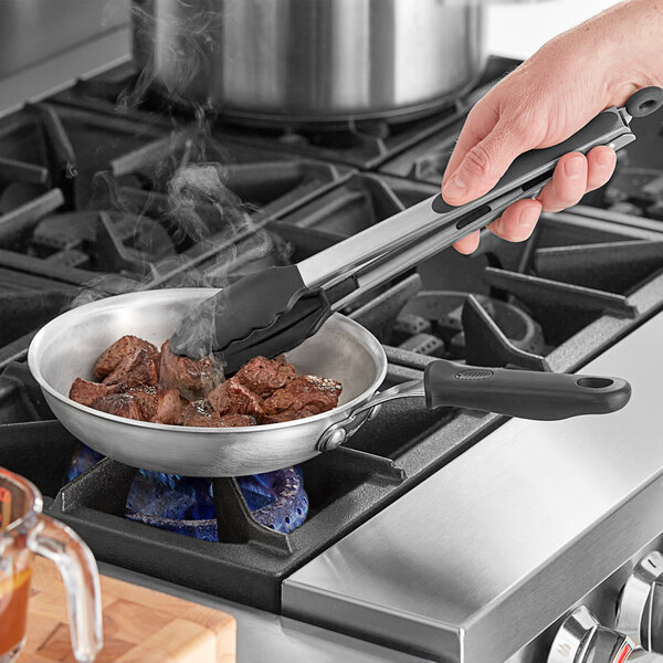 A person using Vollrath Wear-Ever skillet and tongs to cook meat on a stove.